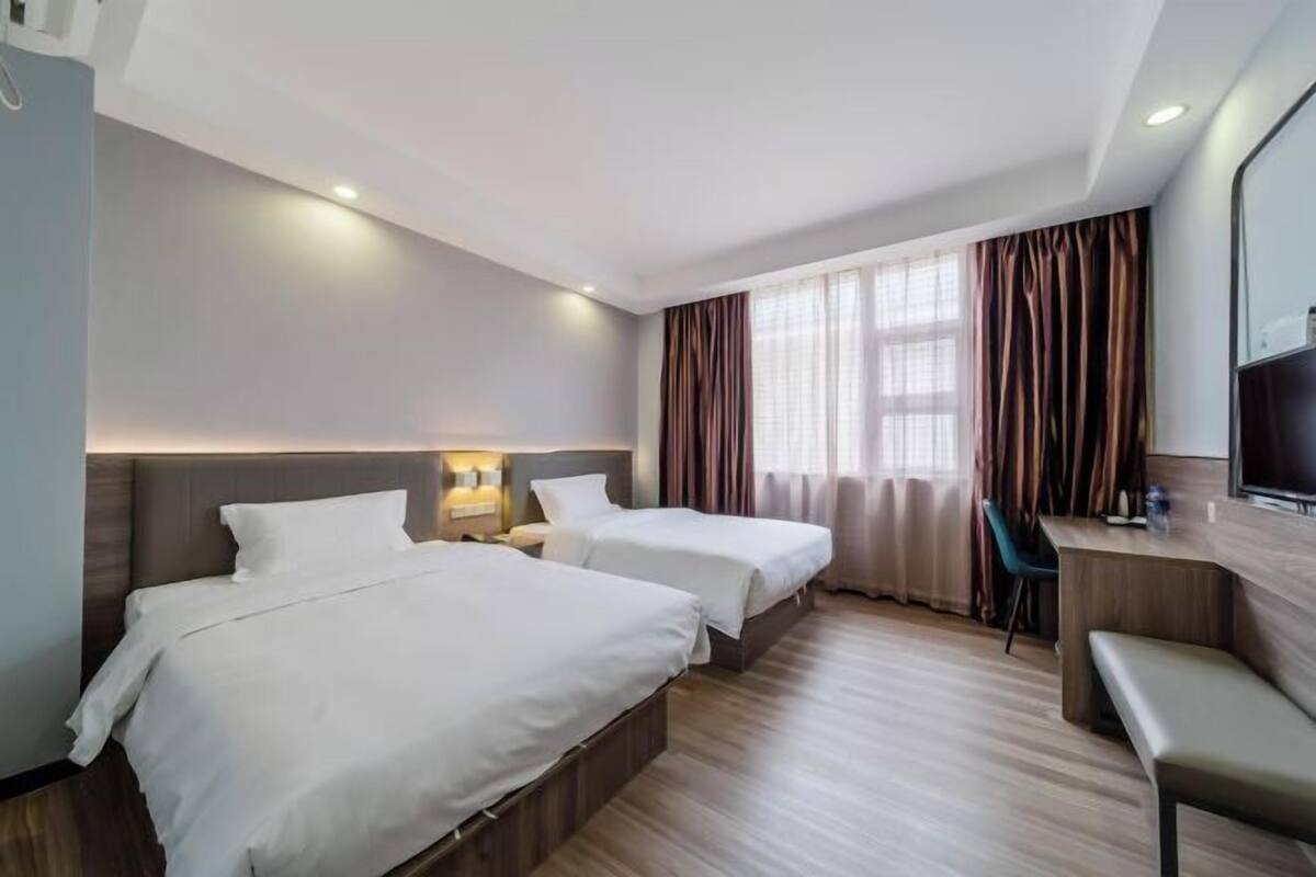 Quarto de hotel com duas camas, uma escrivaninha, uma cadeira, uma televisão e uma janela com cortinas.