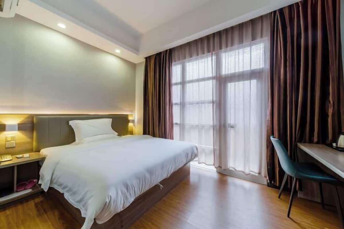 Quarto de hotel com cama, mesa de cabeceira, luminária, cadeira e uma grande janela com cortinas.