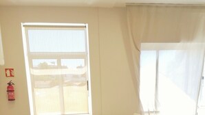 Une pièce avec une table et des chaises blanches, un canapé, une fenêtre avec des stores et un extincteur accroché au mur.