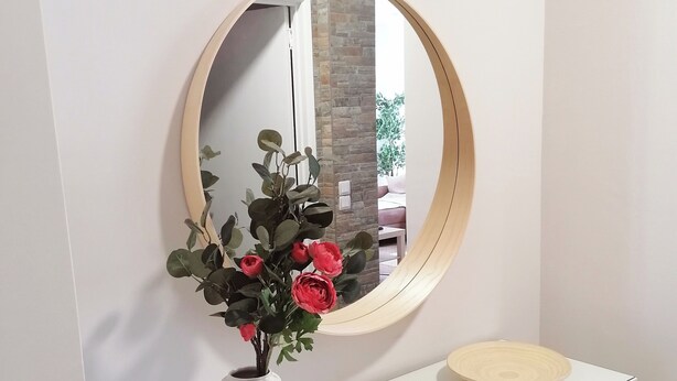 Une console blanche surmontée d’un miroir rond, un vase blanc contenant des feuilles vertes et des fleurs rouges, et un plateau en bois posé sur la console.