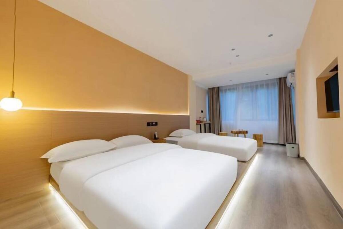 Une chambre d’hôtel avec deux lits, une table de chevet avec une lampe, une grande fenêtre avec des rideaux et un téléviseur à écran plat fixé au mur.