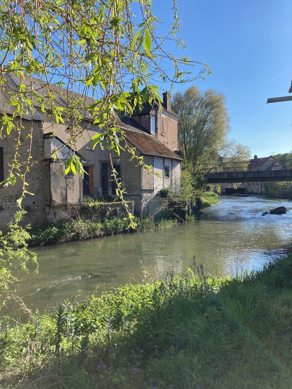 A rustic stone building with a river in front, a bridge in the background, and lush greenery.