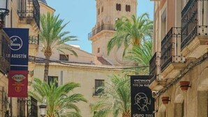 A street with outdoor seating, palm trees, and a tower in the background. - Studio Glamour Rose Alicante (Alicante)