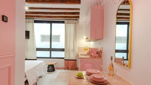A bedroom with a bed, a small table with a chair, a window with curtains, and a kitchenette area with pink appliances. - Studio Glamour Rose Alicante (Alicante)