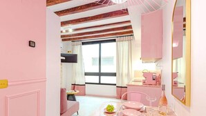 A pink-themed room with a dining table, chairs, a sofa, a small table with a chair, and a window with curtains. - Studio Glamour Rose Alicante (Alicante)