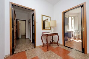 Um corredor com um espelho, uma mesa com abajur, e vista para uma cozinha.