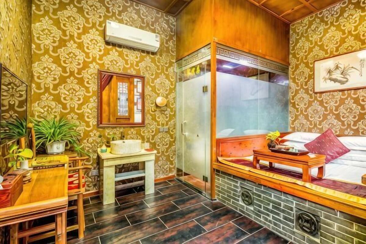 Um quarto com papel de parede estampado, um banco de madeira, uma mesa pequena, um espelho e uma janela.
