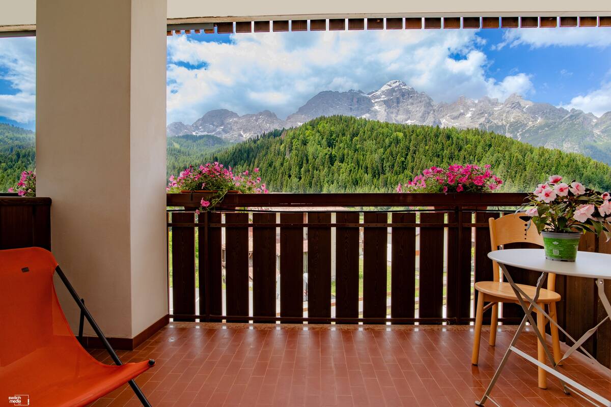 A balcony with a view of mountains, a wooden fence, a table with a chair, and a potted plant.