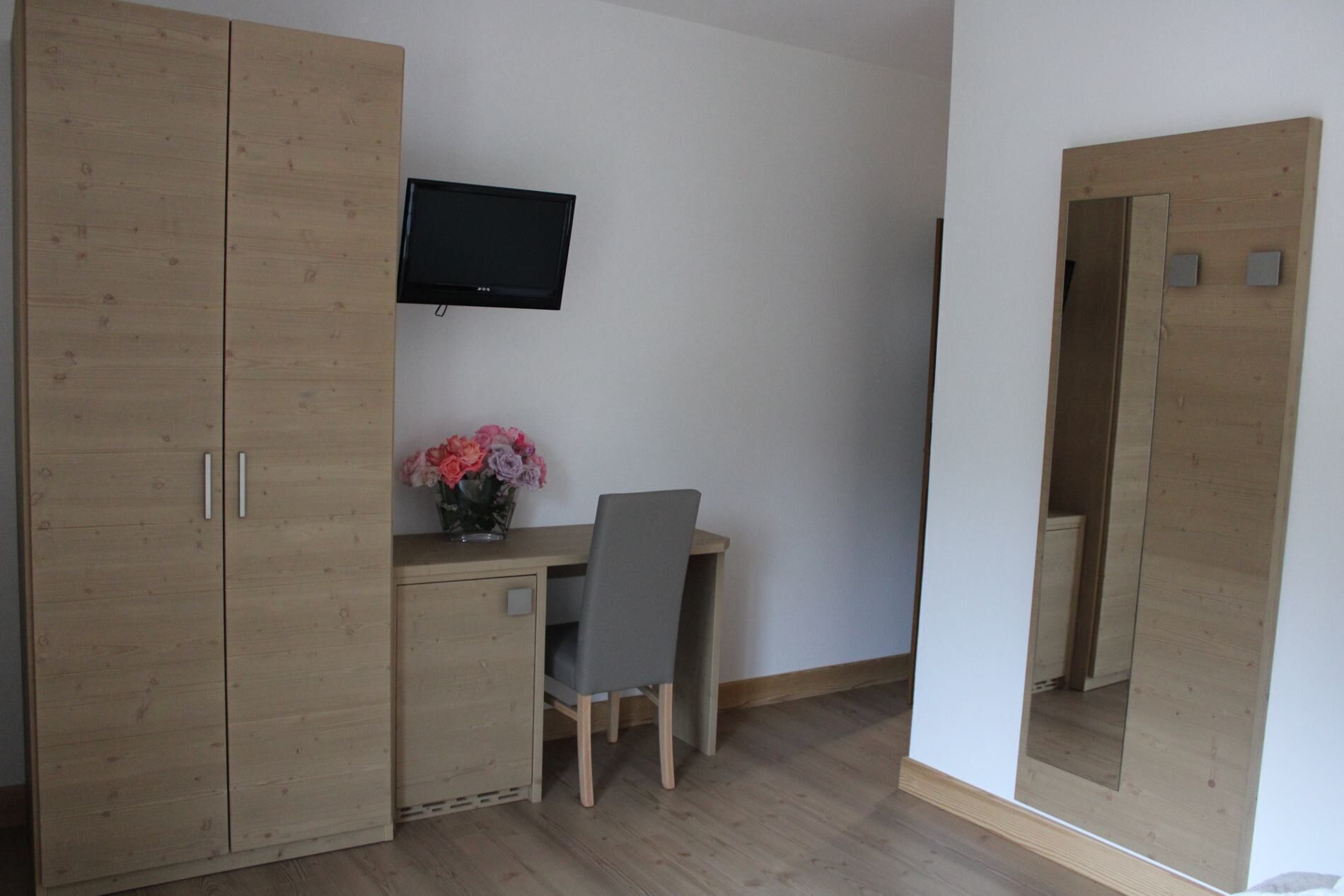 A room with a wooden wardrobe, a desk with a chair, a TV mounted on the wall, and a mirror on the wardrobe door.