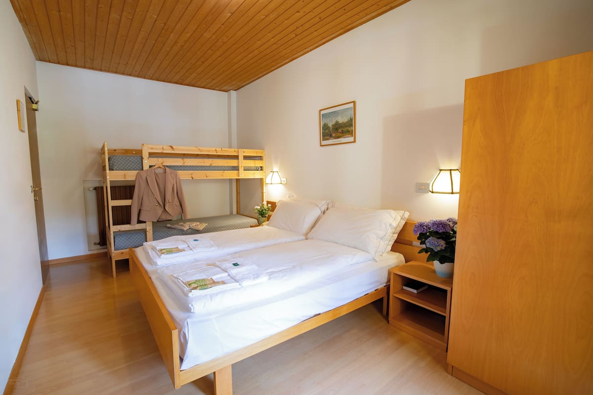 A bedroom with a bunk bed, a single bed, wooden flooring, a wooden ceiling, a nightstand with a lamp, and a wall-mounted shelf.