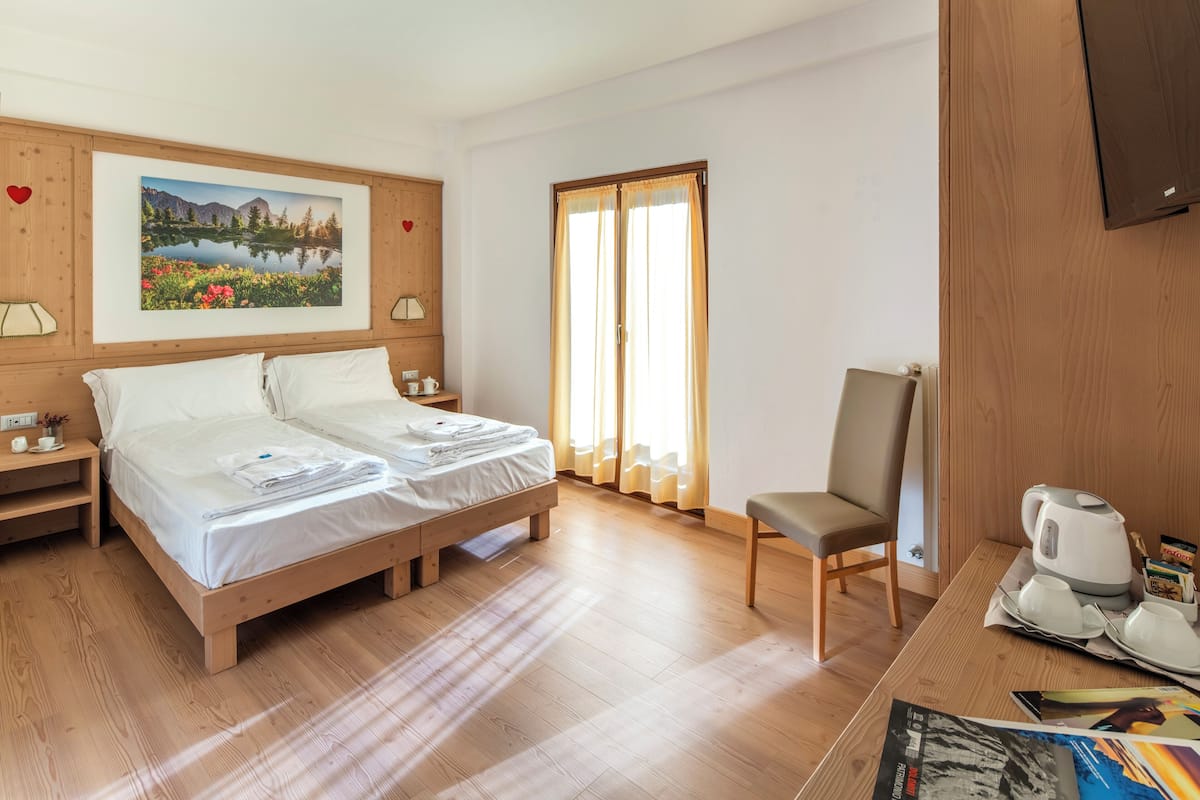 A hotel room with a wooden bed, a chair, a table with a kettle and cups, and a painting on the wall.