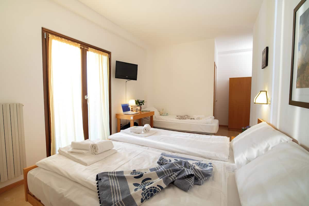 A hotel room with two beds, a desk with a laptop, a TV on the wall, and a door leading to another room.