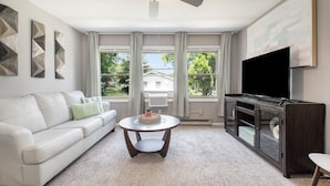 A living room with a white sofa, a round coffee table, a TV stand, and large windows with curtains.