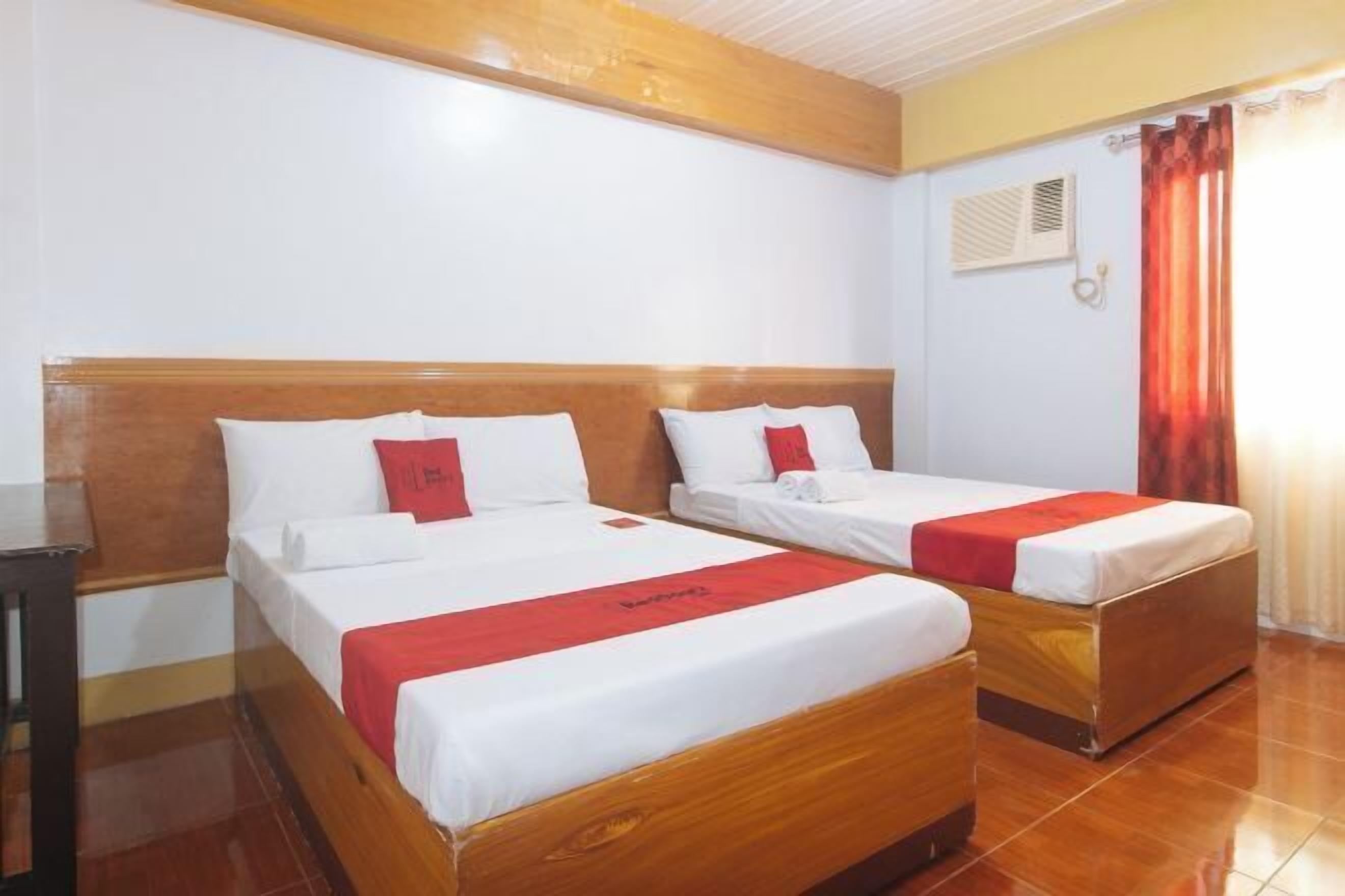 A hotel room with two wooden beds, white bedding, red accents, a wooden headboard, a small table, and a window with curtains.