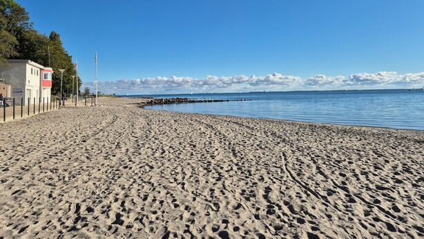 A beach with a sandy shore, a pier, and a building with a red and white facade. - Mobys Hafenglück - Gemütliche 3-zimmer-wohnung für Groß und Klein (Neustadt in Holstein)