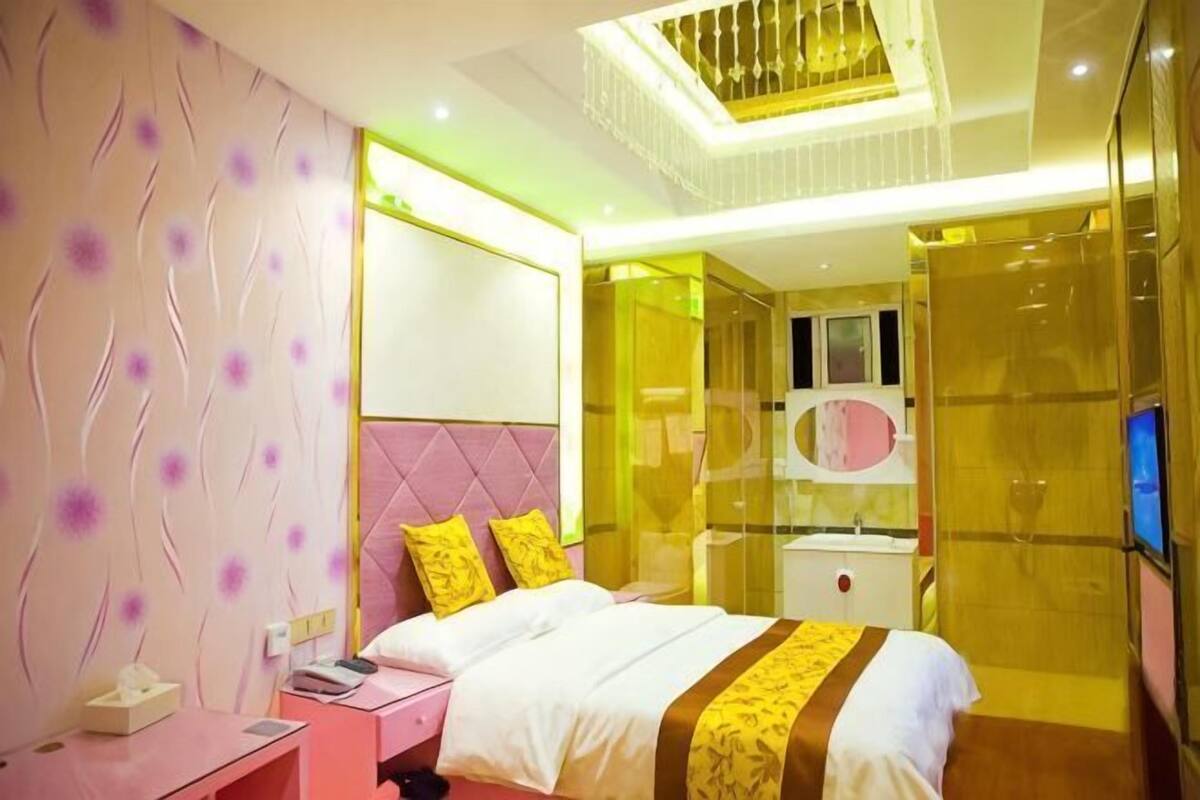 Une chambre d’hôtel moderne aux couleurs rose et blanc, avec un lit recouvert d’une courtepointe blanche et marron, une table de chevet et un grand miroir.
