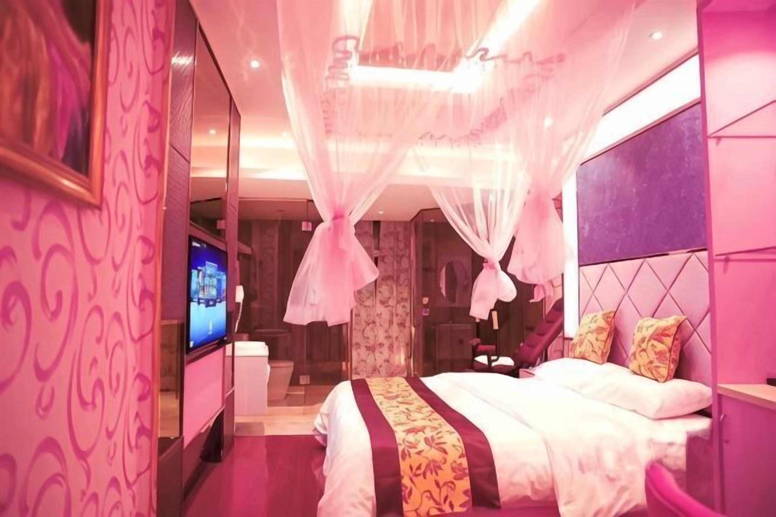 Habitación de hotel decorada en tonos rosados, con una cama, un televisor y un espejo.
