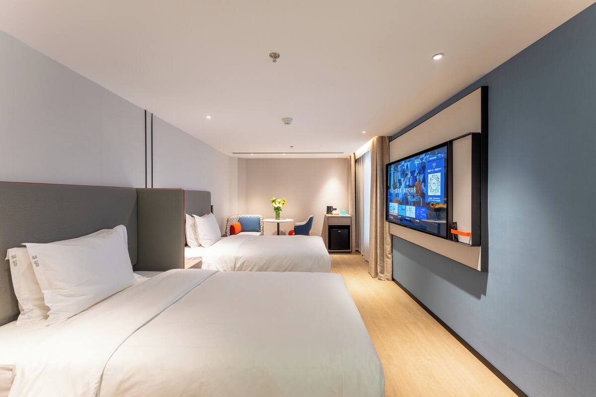 A hotel room with two beds, a TV mounted on the wall, a desk with a chair, and a small table with a vase of flowers.