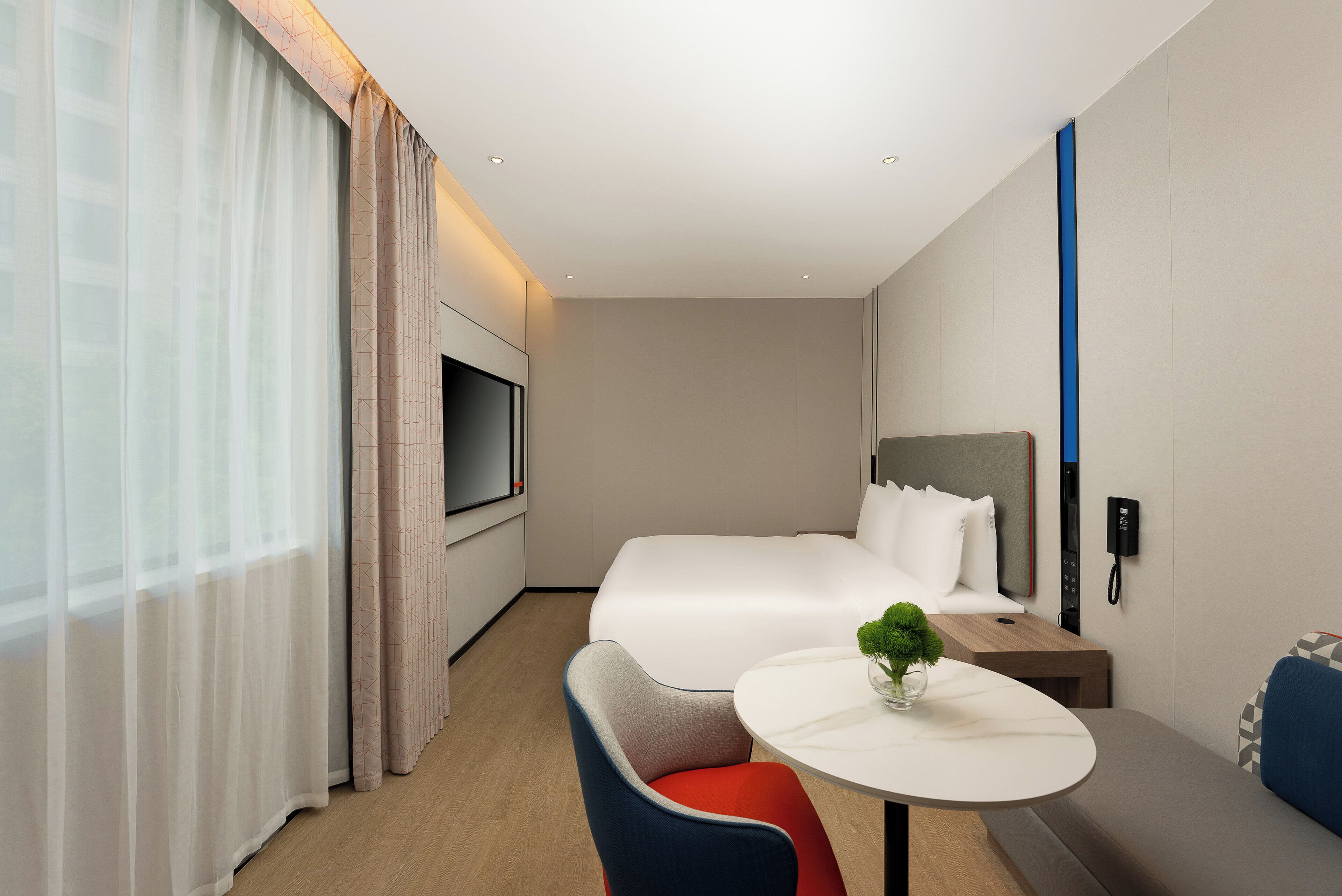 Una habitación de hotel moderna con cama, televisión, mesita de noche con una planta, una silla y una pequeña mesa.