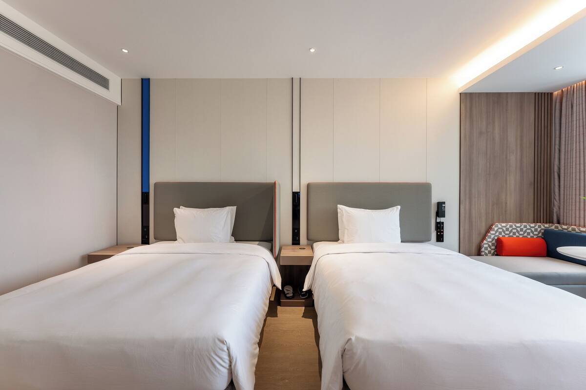 A hotel room with two beds, a small table, and a seating area with a checkered cushion.