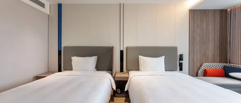 A hotel room with two beds, a small table, and a seating area with a checkered cushion.