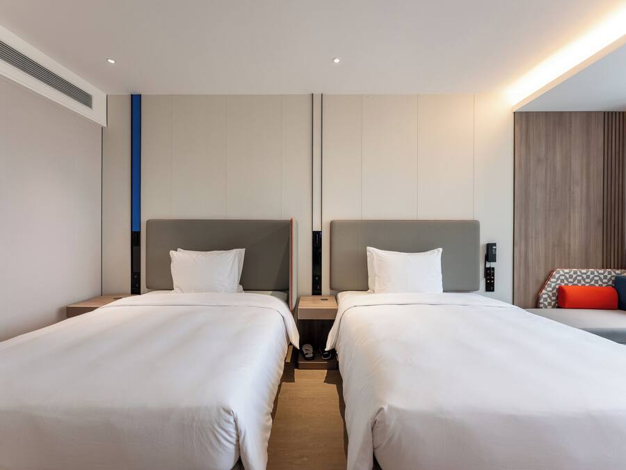 Habitación de hotel con dos camas, una mesita pequeña y un rincón de descanso con cojín a cuadros.