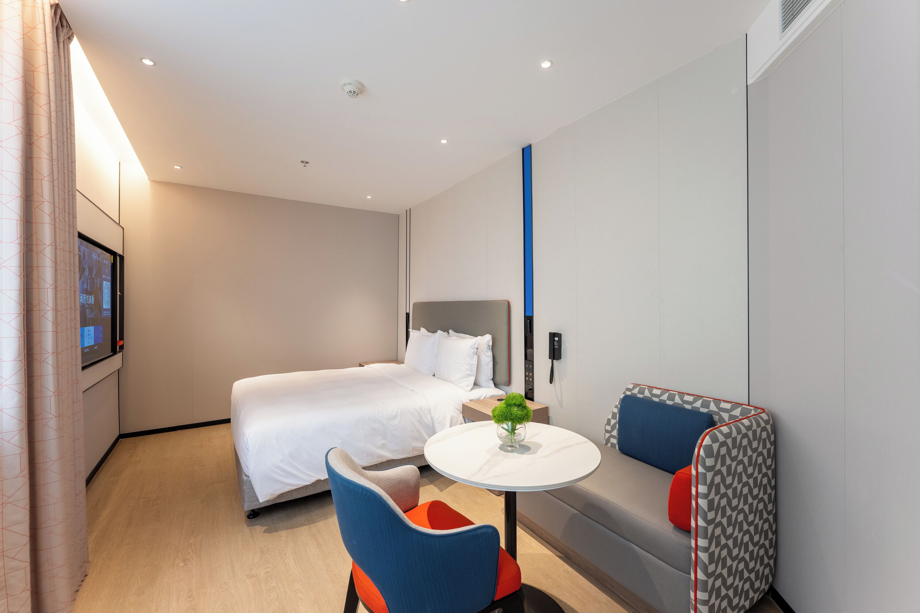 Una habitación de hotel moderna con una cama, una pequeña mesa redonda con un florero, un sofá con cojines y un televisor montado en la pared.