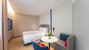 Una habitación de hotel moderna con una cama, una pequeña mesa redonda con un florero, un sofá con cojines y un televisor montado en la pared.