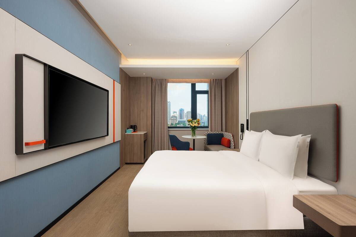A modern hotel room with a large bed, a flat-screen TV mounted on the wall, a small desk with a chair, and a city view through the window.