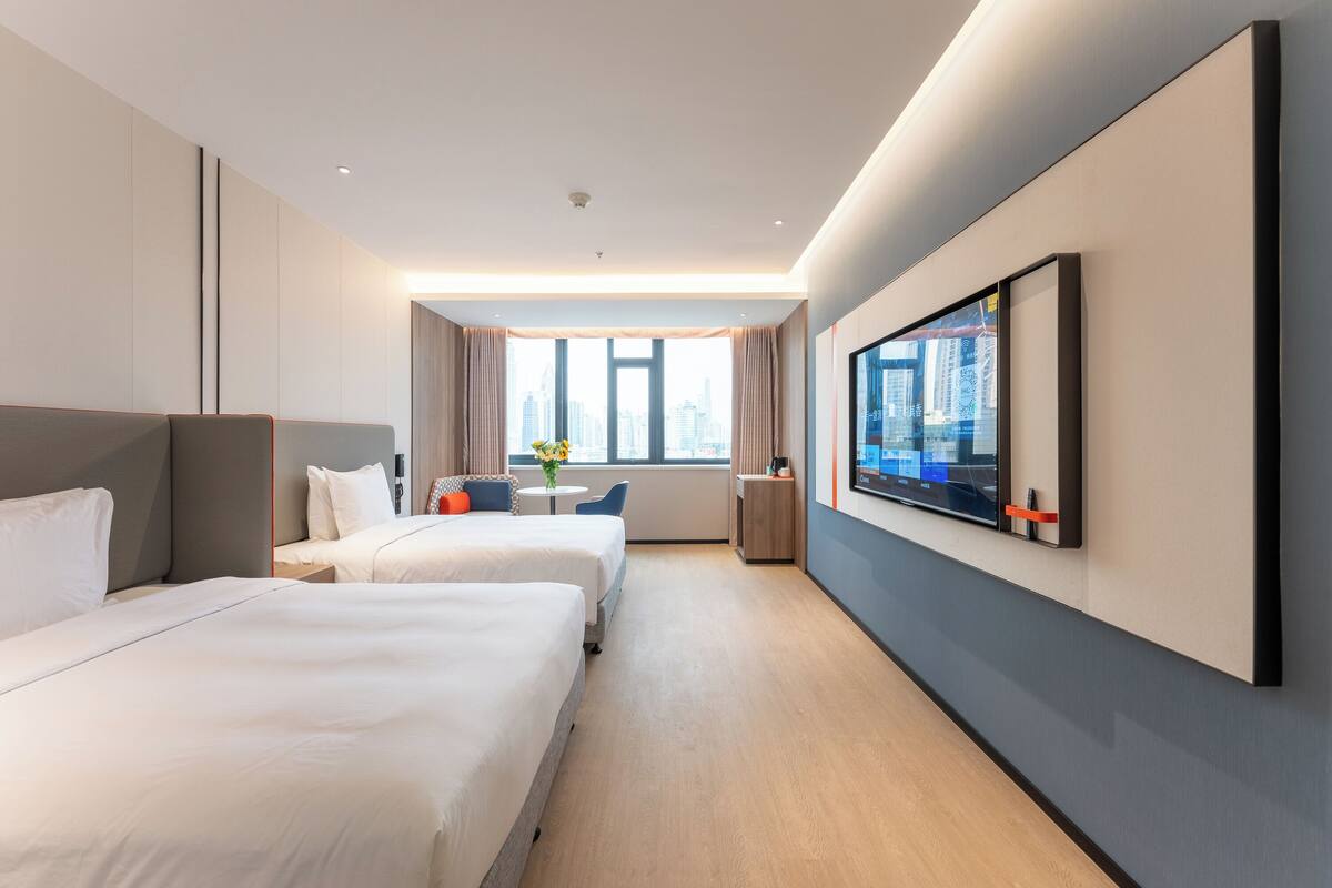 A modern hotel room with two beds, a large TV, and a city view.