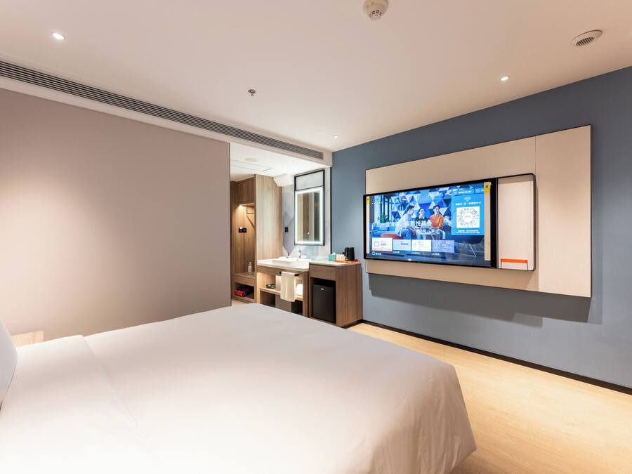 Una habitación de hotel moderna con una cama grande, un televisor montado en la pared, un baño con lavabo y espejo, y un vestidor.