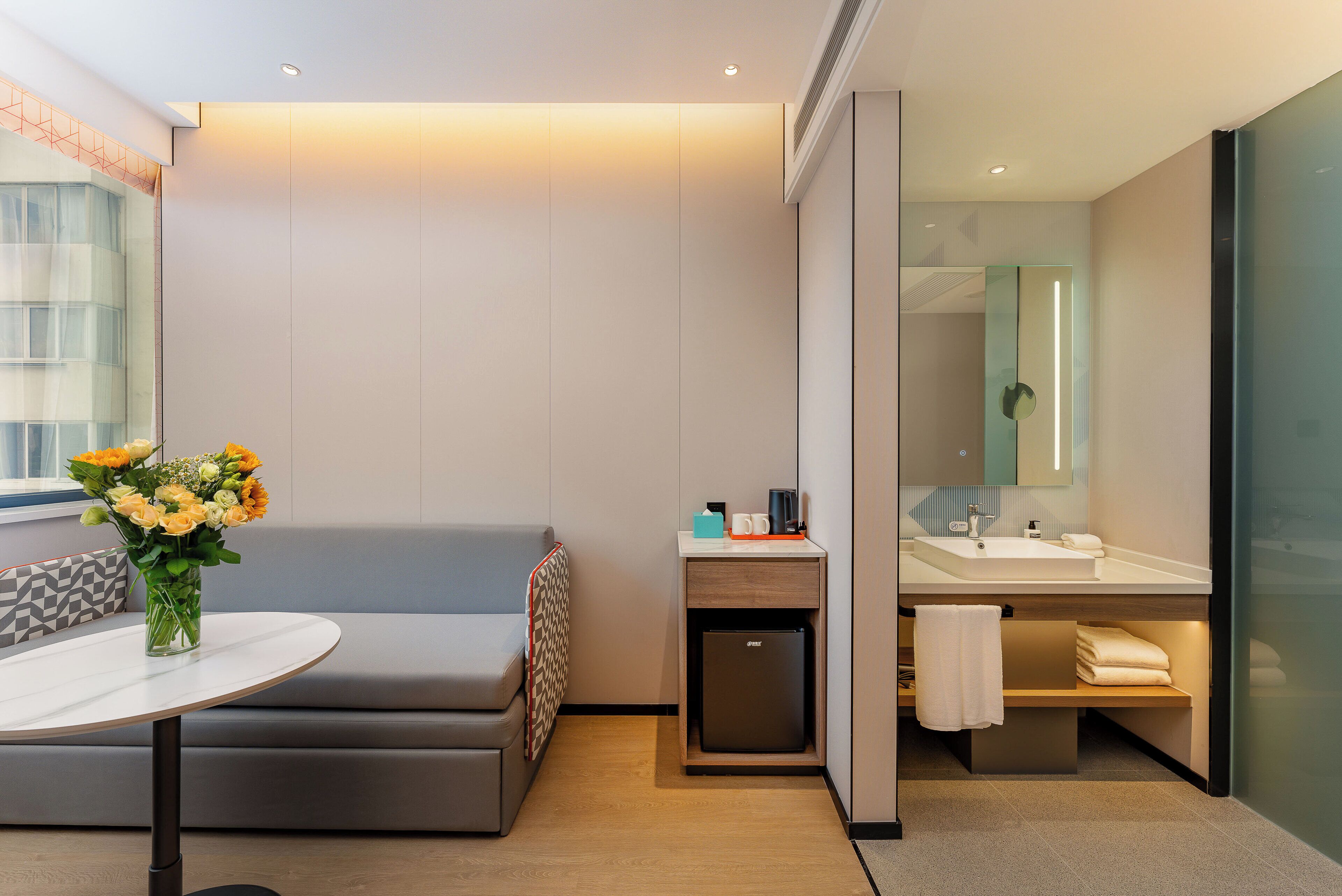 Una habitación de hotel moderna con un sofá, una pequeña mesa con un jarrón de flores, un refrigerador compacto, un lavamanos y un espejo.