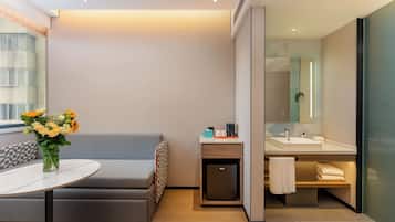 Una habitación de hotel moderna con un sofá, una pequeña mesa con un jarrón de flores, un refrigerador compacto, un lavamanos y un espejo.