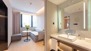 Habitació d'hotel moderna amb un sofà, una taula rodona amb flors, un mirall gran i un bany amb un lavabo i barra de tovalloles.