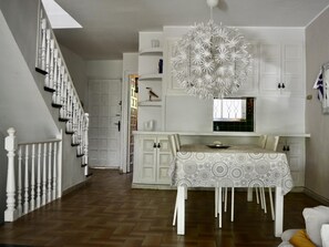 Un coin repas avec une nappe à motifs, des meubles blancs, un escalier blanc et un luminaire décoratif au plafond.
