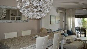 Een eethoek met een tafelkleed met patroon, witte stoelen, een decoratieve plafondlamp en een bank met kussens.