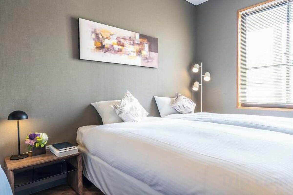 A hotel room with a bed, bedside table, lamp, and a window with blinds.