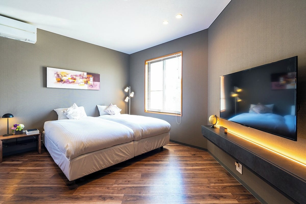 A modern bedroom with a large bed, a flat-screen TV mounted on the wall, a window with blinds, and a bedside table with a lamp and a plant.