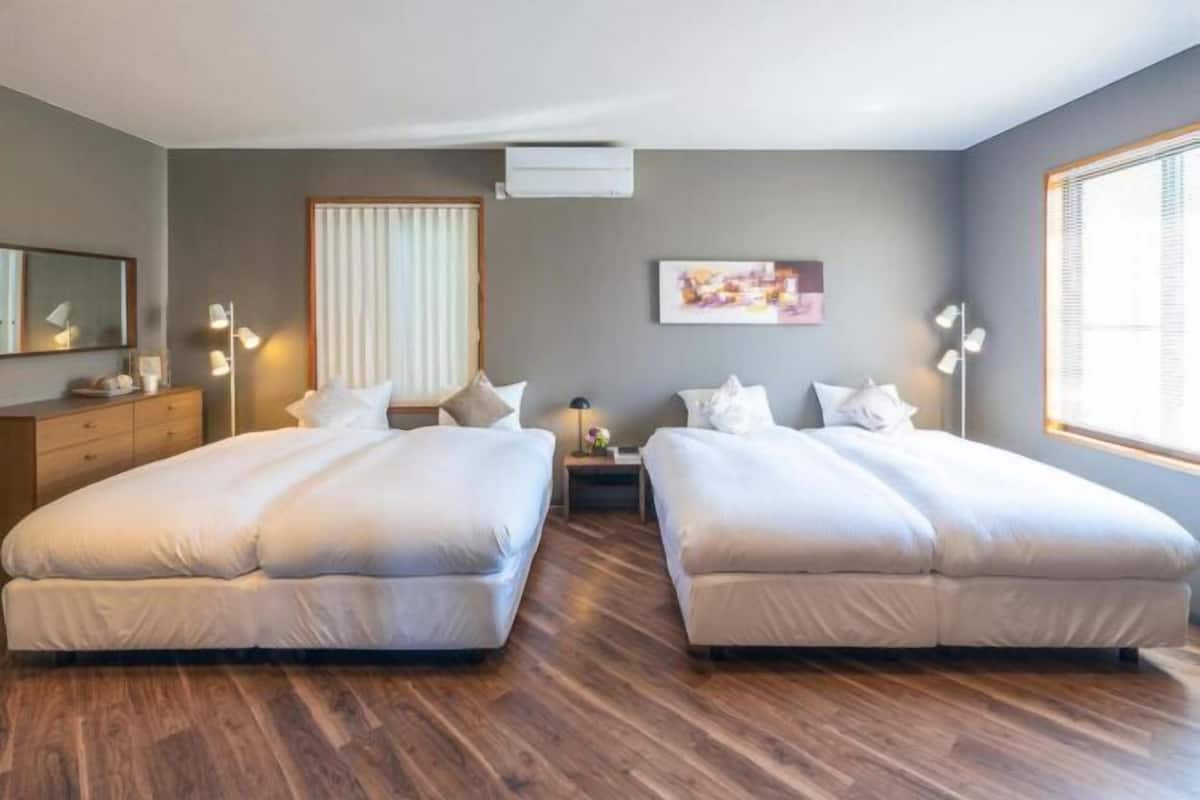 A hotel room with two beds, a wooden floor, a window with blinds, a bedside table with a lamp, and a wall-mounted air conditioner.