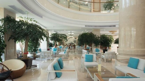 Eine geräumige Lobby mit mehreren Sitzbereichen, großen Kübelpflanzen und einer hohen Decke mit einem zentralen Kronleuchter.