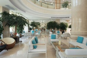 Eine geräumige Lobby mit mehreren Sitzbereichen, großen Kübelpflanzen und einer hohen Decke mit einem zentralen Kronleuchter.