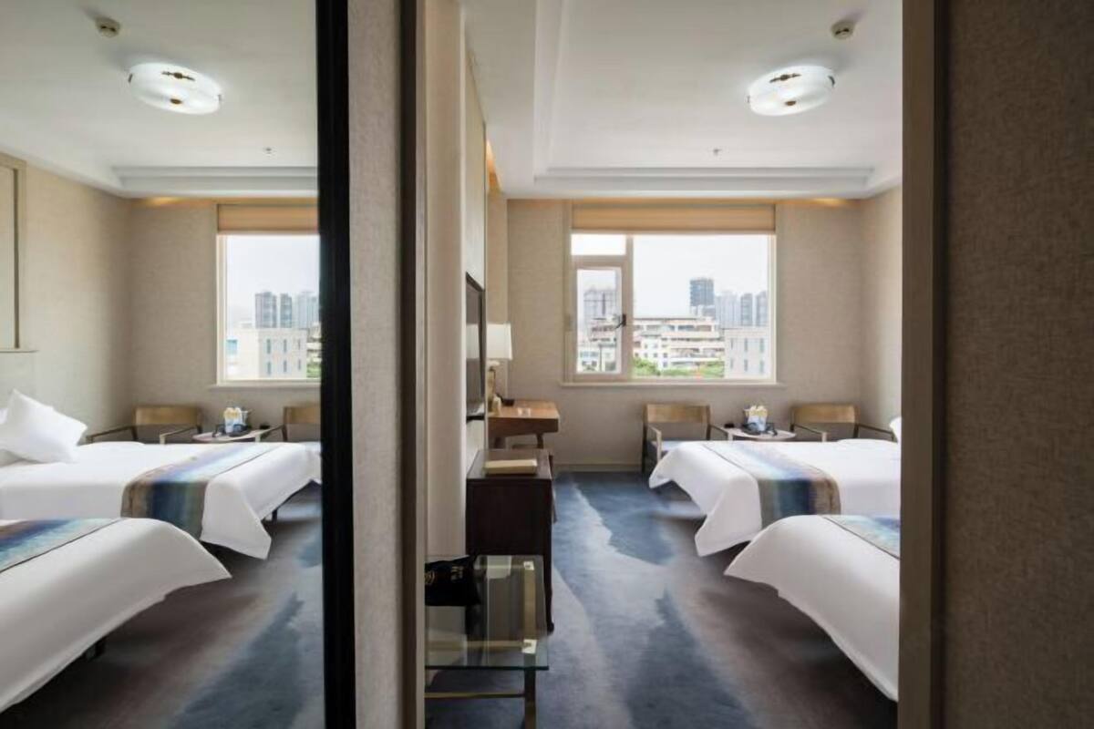 A hotel room with two beds, a desk, a chair, a TV, and a large window with a city view.