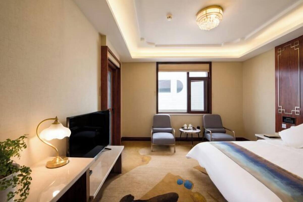 A hotel room with a bed, a desk with a lamp, a TV, two chairs, a small table with cups, and a window.