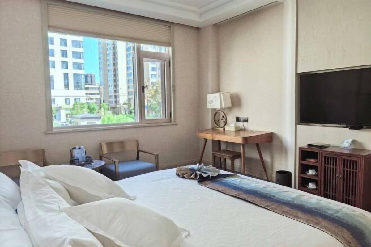 A hotel room with a bed, a desk, a chair, a TV, and a window with a view of buildings.