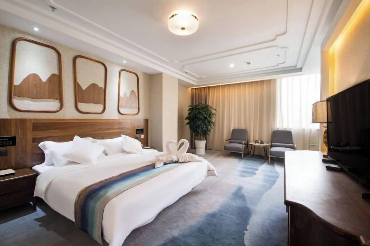 A hotel room with a bed, a desk, a TV, a plant, and artwork on the wall.