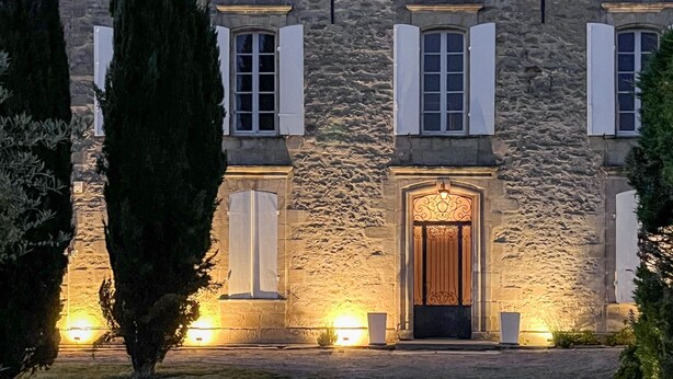 Un edificio de piedra con una entrada central, flanqueada por ventanas con contraventanas, e iluminada por focos en el suelo.