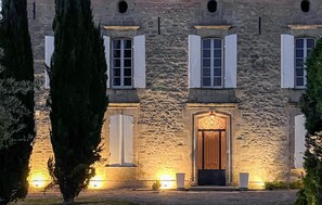 A stone building with a central entrance, flanked by windows with shutters, and illuminated by ground lights.