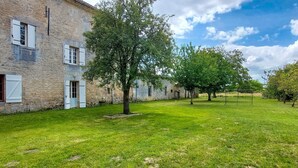 Un edificio de piedra con contraventanas blancas, un patio cubierto de césped y árboles.