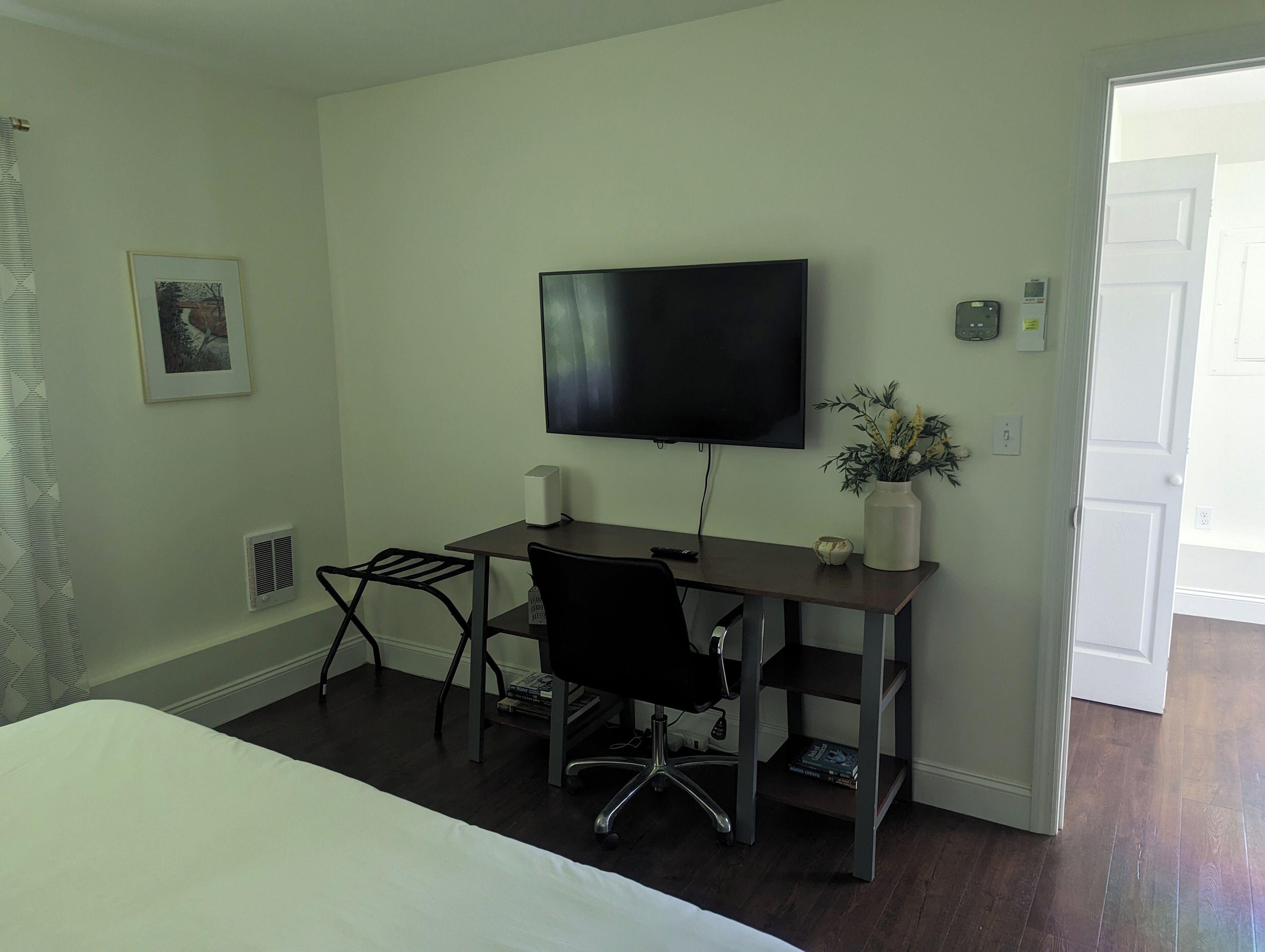 Habitación de hotel con cama, escritorio con computadora y altavoces, silla, televisor, planta pequeña y puerta.