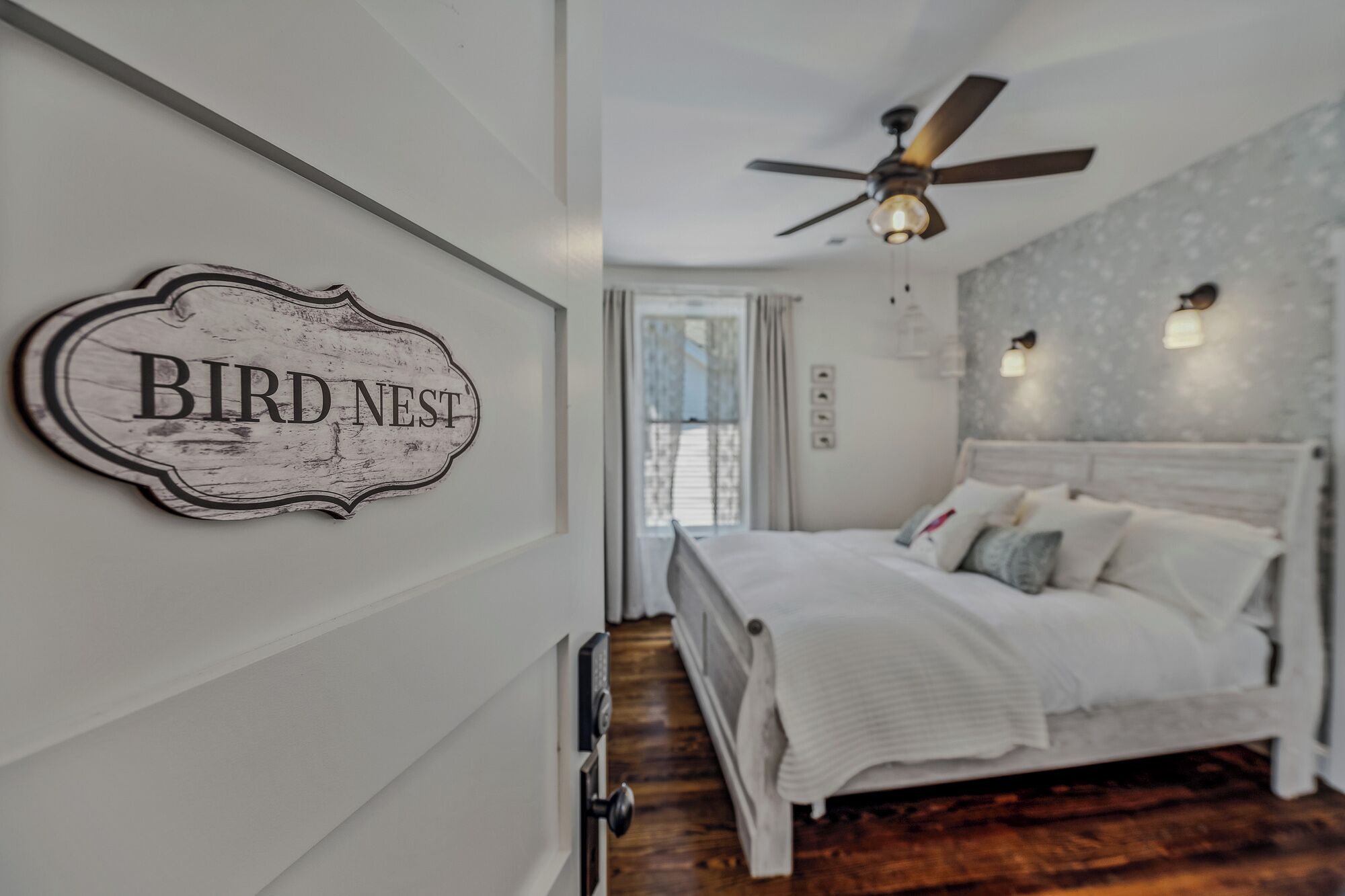 A bedroom with a bed, a ceiling fan, a wall-mounted sign reading "BIRD NEST", and a window with curtains.
